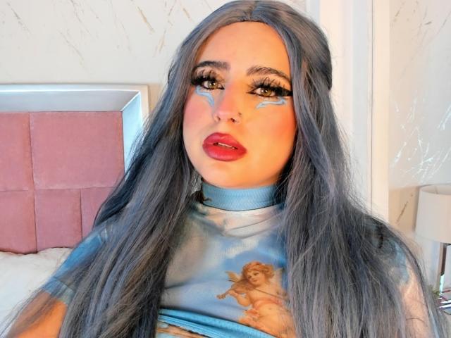 StephanieMoretti with long gray hair, blue face paint, red lipstick, and a blue shirt with a cherub print, showing her tits slightly, in a bright room.