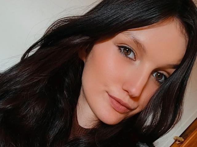 AdelineCreazy has dark, wavy hair, light skin, and brown eyes. She's smiling with subtle makeup, showing her cute nose and full lips. Her tits are not visible.