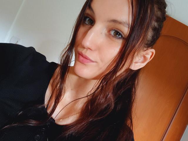 AdelineCreazy has long brown hair, green eyes, and a slight smile. She's wearing a black top, showing her bare shoulders and a hint of her cleavage.
