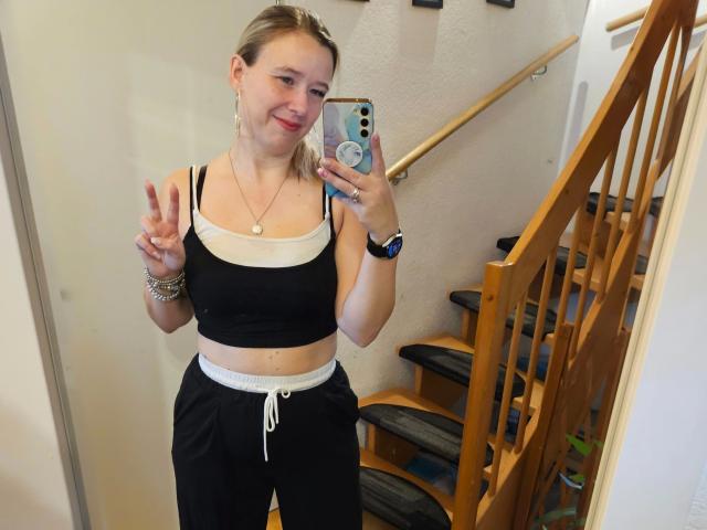 Lea-sommer-hot with blonde hair, smiling, wearing black sports bra and black pants, flashing a peace sign, holding phone with blue case, standing in front of wooden staircase.