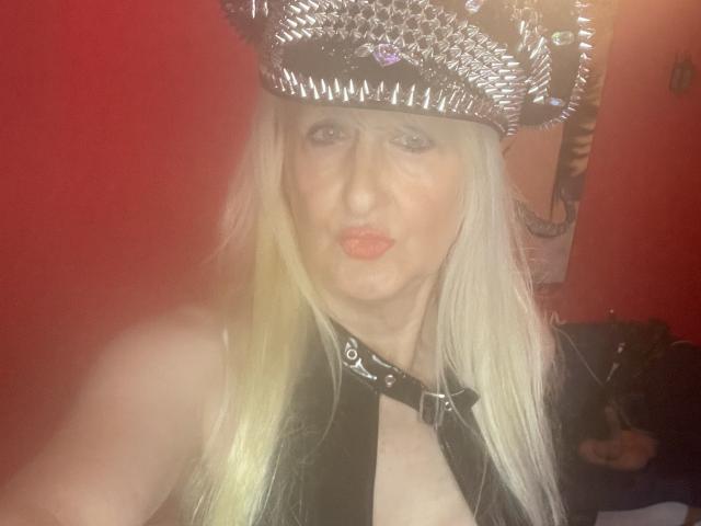 LadyHazel, con cabello rubio largo, vistiendo un sombrero de metal con púas y un collar de cuero negro, haciendo pucheros con labios rojos, mostrando sus hombros desnudos y una blusa negra en una habitación iluminada con luz roja..