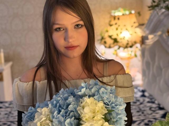 Bubochka with long brown hair, off-shoulder gray top, holding a bouquet of blue and white hydrangeas, looking directly at the camera with a subtle smile.