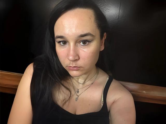 SaliBerry has pale skin, black hair, and is looking down with a serious expression. She's wearing a black tank top, showing her small tits, and has a necklace.