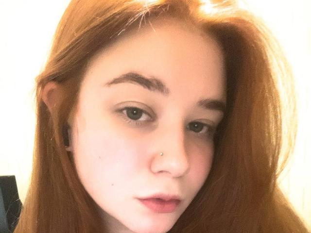EmmaLands tem pele clara, cabelo ruivo, olhos castanhos, um piercing no nariz e alargadores de orelha pretos, com um leve franzido nos lábios rosados, olhando suavemente para a câmera..
