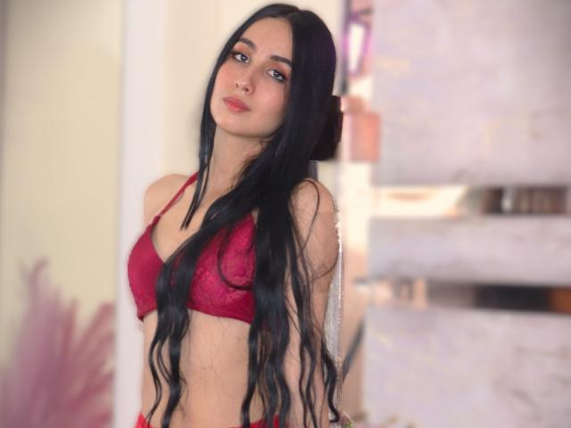ValenStorm with long black hair, wearing a red lace bra, showing her small tits, standing with a slight smile, looking at the camera. Soft-focused background.