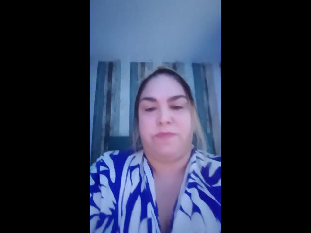 ChubbyAngel69 has a chubby face with light skin, brown eyes, and blonde hair, wearing a blue and white floral shirt, looking down with a slight smile.
