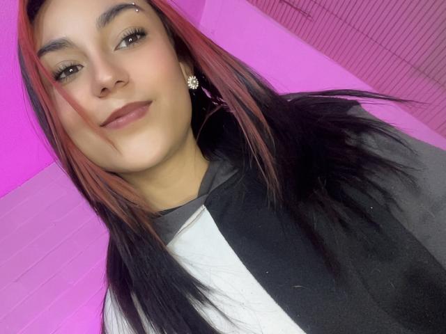 AmyStreetwise69 has red and black hair, brown eyes, a nose piercing, and is smiling with a subtle makeup look. She's wearing a black and white shirt with star earrings.