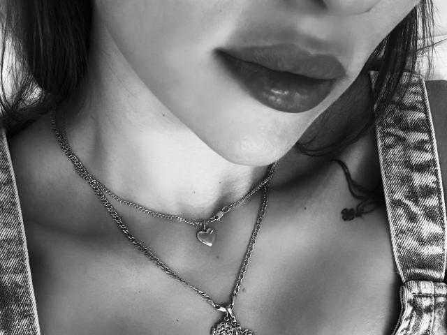 Il viso di TheFatalLady, con labbra piene e rossetto scuro, indossa una collana a forma di cuore e ha un piccolo tatuaggio sul lato destro. Sono visibili le clavicole e le bretelle in denim..