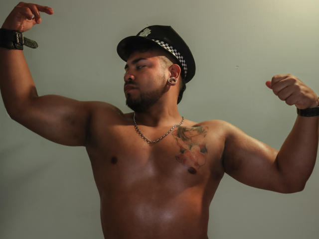 BrianMarly, shirtless, flexing biceps, wearing black police hat, chain necklace, tattoo on right chest, black wristbands, earbowl, short beard, eyes closed, muscular, medium brown skin, light green wall.