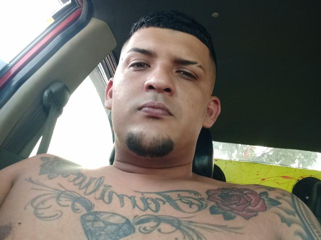 JorweenHot, shirtless, has tattoos on his chest, including "Chismis" and a rose, short black hair, and a goatee, sitting in a car, looking down.