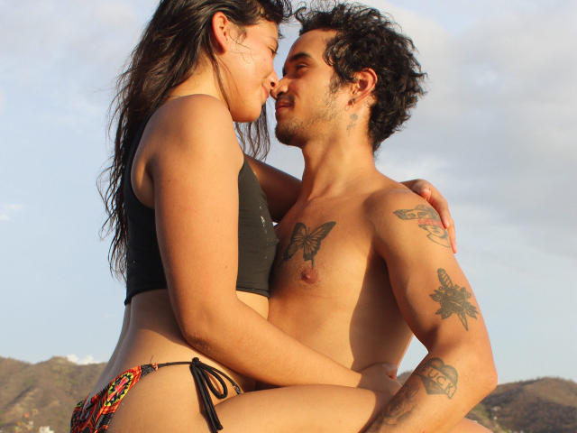 LailaAsher with dark hair and black top, sitting on a tattooed man with curly hair, both shirtless, kissing, her ass near his crotch, colorful bikini bottom, sunny.