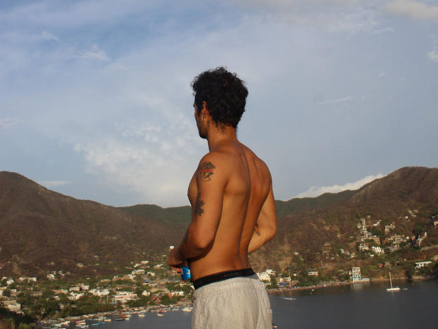 Shirtless LailaAsher with curly black hair, tan skin, and tattoos on his right shoulder, wearing gray shorts, stands holding a blue bottle, facing a coastal town and mountain.