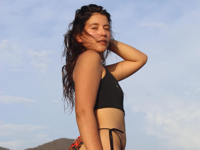LailaAsher with wet, dark hair, wearing a black sports bra and colorful bikini bottoms, hand in hair, side profile, blue sky, and distant mountain. Her tits are small.