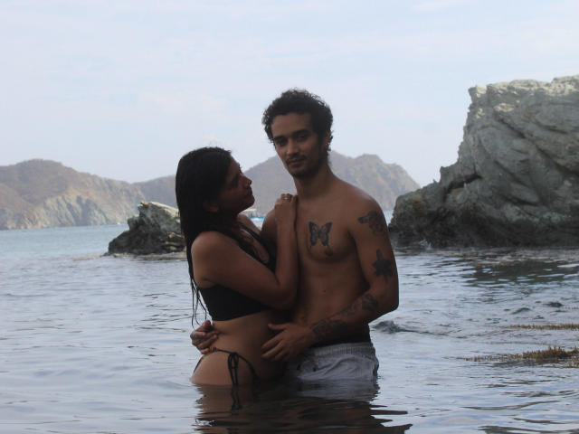 LailaAsher with long black hair and small tits, wearing a black bikini bottom, standing in water, embracing a tattooed, shirtless guy with a butterfly tattoo on his chest.