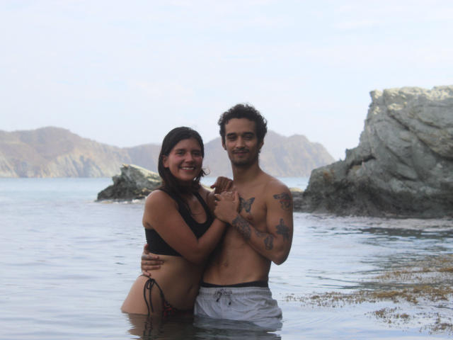 LailaAsher, com cabelo castanho e biquíni preto, está em águas rasas, com os braços ao redor de LailaAsher, que está sem camisa, com tatuagens e shorts cinzas, e ambos estão sorrindo. Pedras ao fundo..