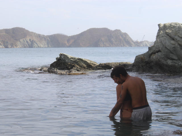 LailaAsher stands in shallow water, shirtless, with a tattoo on his right arm, wearing gray shorts, looking down at his hands, with rocky cliffs in the background.