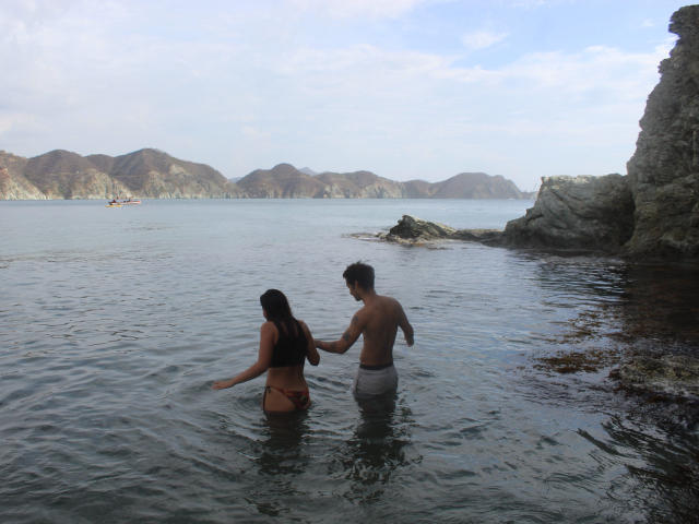 LailaAsher with long black hair in a bikini stands in shallow water, touching a shirtless guy with short brown hair in white pants, near rocky shoreline.