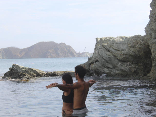 Laila Asher e un uomo senza camicia con un afro riccio sono in piedi in acque poco profonde, con le braccia tese l'uno verso l'altro, con scogliere rocciose e una montagna in lontananza sullo sfondo..