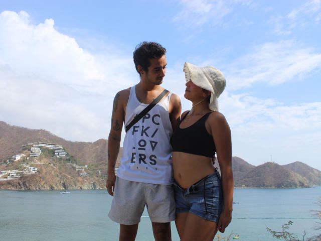 Laila Asher è in piedi accanto a un uomo alto, la cui canotta bianca e i pantaloncini grigi mostrano la scritta "FK/97 ERS". Lei indossa un reggiseno sportivo nero e pantaloncini di jeans, ed ha un cappello da sole..