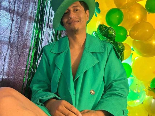 SandyAndMike in a green hat and coat, smiling with a small mustache, sitting among green and yellow balloons. His ass is barely visible in the bottom left.