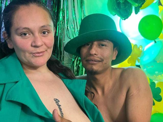 SandyAndMike with a green top and a tattoo, and Mike shirtless in a green hat, both smiling, with green and yellow balloons in the background.