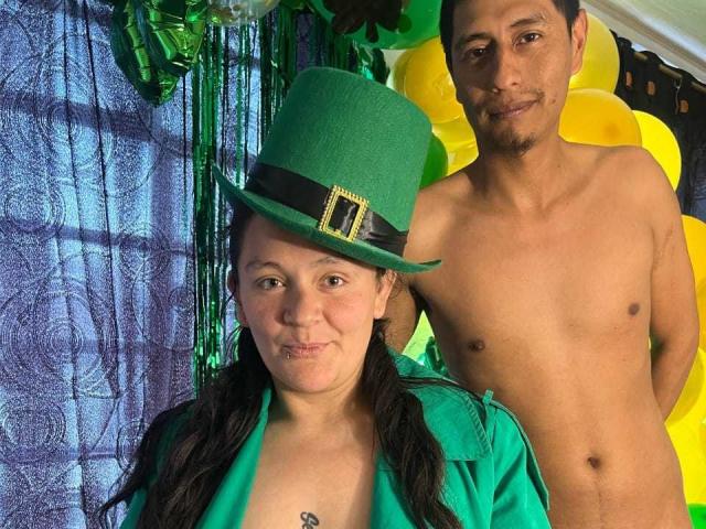 SandyAndMike wearing a green top hat and green shirt, with pigtails, stands next to shirtless Mike, both with tan skin, in a festive green and yellow setting.