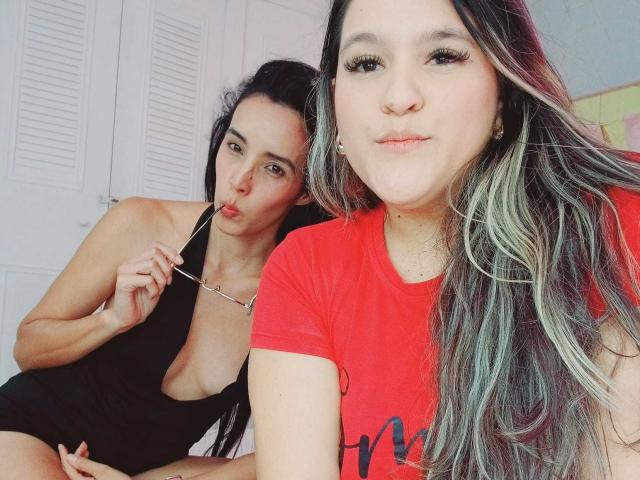 JenniferAndMia with long black and blonde hair in red shirt, pouty lips, and small nose, sitting next to Mia with black dress, showing off her big tits, sucking on glasses.