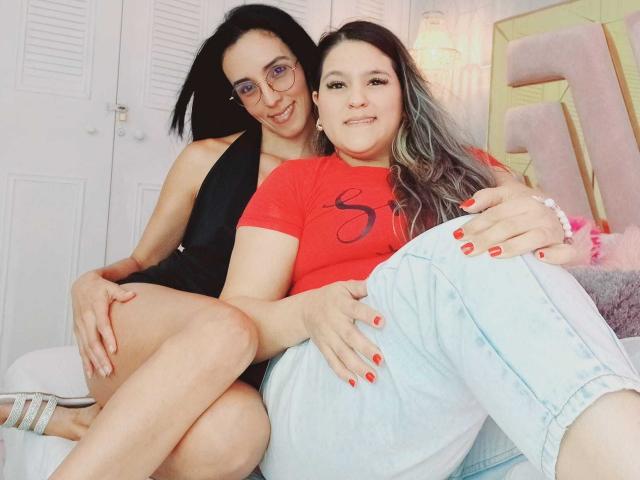 JenniferAndMia with black hair in a black dress and glasses, sitting on JenniferAndMia with gray hair in a red shirt and light jeans, showing red nails.