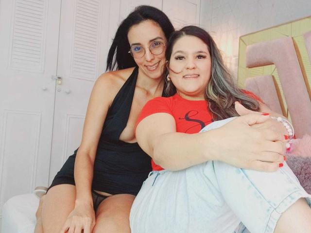 JenniferAndMia with black hair and glasses, wearing a low-cut black dress, sitting next to Mia with long brown hair, red shirt, and blue jeans, showing her tits.