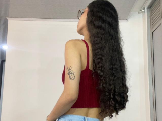 ClarelandsWell with long curly black hair, red crop top, light blue jeans, tattoo on upper right arm, looking over shoulder, wearing glasses, standing in a room.