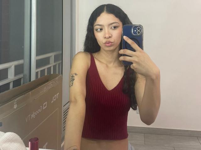 ClarelandsWell with long black hair, red ribbed tank top, and small tits, taking a selfie in a mirror, pouting lips, holding a blue phone, with a tattoo on her arm.