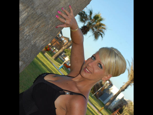 MistressCathy-hot with blonde hair, smiling, leaning against a tree, wearing a black top with cutout, showing her boobs, pink nails, and a tattoo on her right chest.