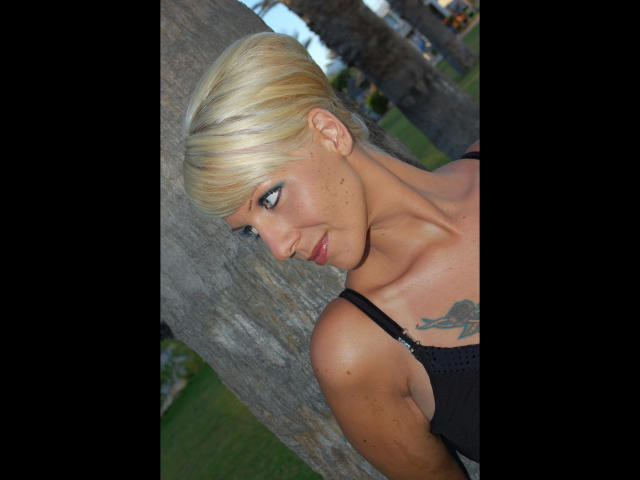 MistressCathy-hot with blonde updo, blue eye makeup, black top, tattoo on right shoulder, leaning against tree, smiling sideways, visible collarbone, outdoors.