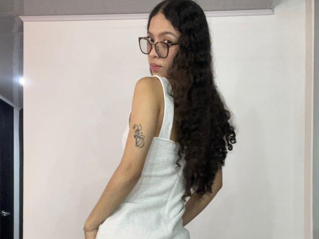 ClarelandsWell with long curly dark hair, glasses, white tank top, and a tattoo on her right arm, looking over her shoulder with a slight smile.