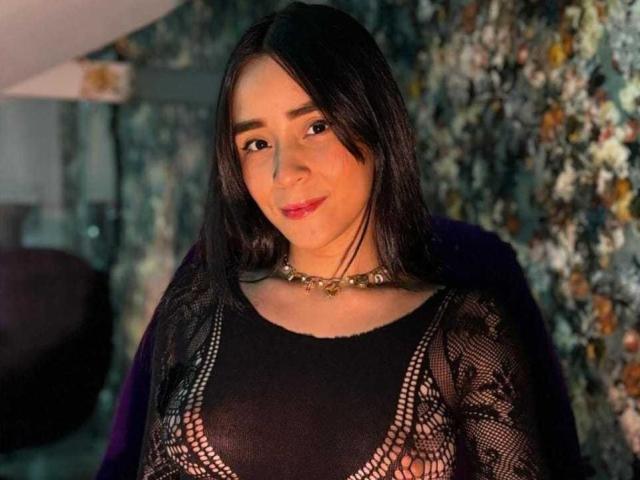 AlissonHall with black hair, wearing a black lace top, showing small tits, and a gold necklace, smiling slightly. Dark, textured background. Warm lighting on her face.