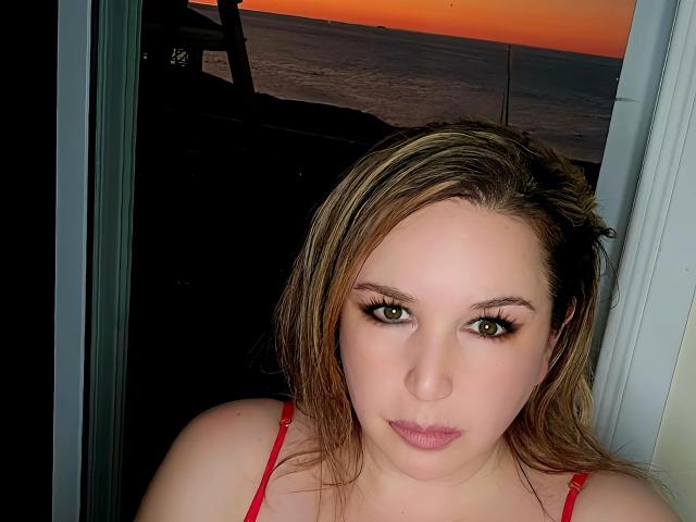 TitanicTits met blond haar, groene ogen en een rode beha, die de camera aankijkt met een neutrale uitdrukking, staand voor een glazen deur tijdens de zonsondergang..