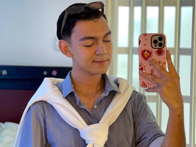 DeusBeauty, short-haired, wearing glasses on head, blue striped shirt, white hoodie, necklace, holding phone with red floral case, smiling, taking selfie, bedroom, daytime.