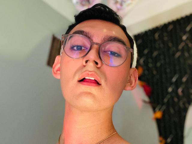 DeusBeauty has short black hair, clear glasses reflecting pink light, slightly open pink lips, and a gold necklace, looking up with a slightly flushed face.