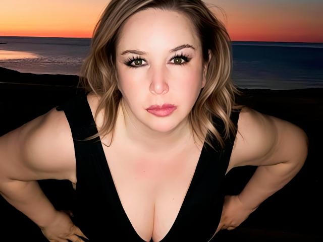 TitanicTits with large boobs, fair skin, and blonde hair, wearing a black dress, hands on hips, looking up with green eyes and pink lips, sunset in the background.