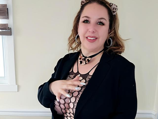 TitanicTits with cat ears, hoop earrings, and a black blazer over a mesh top showing her big tits. She's smiling, pointing with a finger, and has a cute nose.