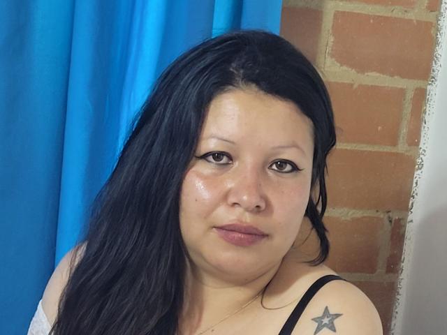 PussyLovePure has black hair, brown eyes, and a star tattoo on her shoulder. She's wearing a black top, with blue curtains and a brick wall behind her.