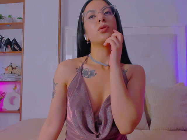 SarahLuna with long black hair, glasses, and a deep purple, shiny, halter-neck dress, pouting with her finger on her cheek, showing tattoos on her chest and arm.