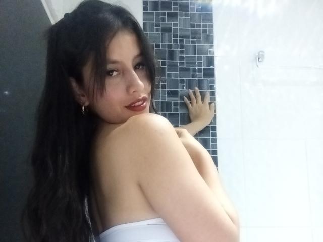 MadissonFontaine with long black hair, red lipstick, and white strapless top, facing camera, showing her back and right boob, hand on tiled wall.