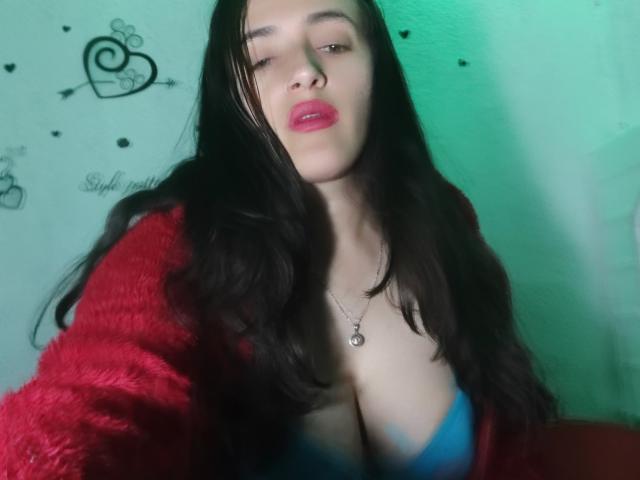 AndreaHotLove69 with long black hair, red fluffy jacket, blue bra, cleavage, red lipstick, green light, necklace, slightly winking, and a sexy, confident expression.