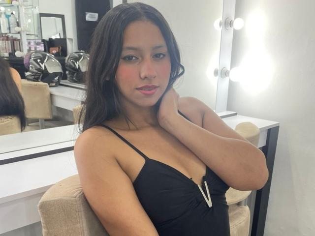 AshleyRosel with long dark hair, wearing a black spaghetti-strap top, showing small tits, pink blush on cheeks, sitting in a bright room with a mirror and lights.