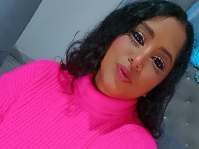 MariaCatalina has dark curly hair, brown eyes with thick eyelashes, and is wearing a bright pink ribbed turtleneck. Her pouty lips are slightly glossy.