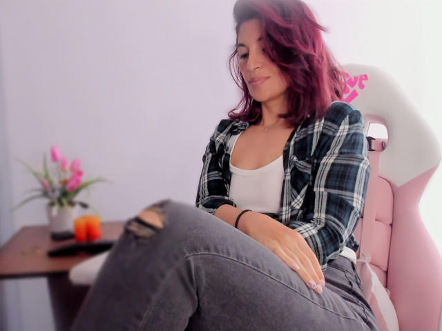 LaurentColin with red wavy hair, white tank top, black plaid shirt, gray ripped jeans, sitting on pink chair, hand on knee, subtle smile, small pink flower on wooden table.