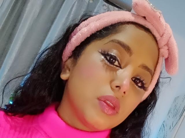 MariaCatalina has tan skin, dark wavy hair, and wears a pink headband with a bow. She has pink lipstick, bold eyeliner, and a pink turtleneck.