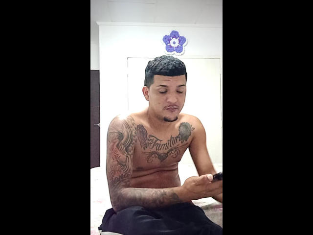 shirtless JorweenHot with tattoos, short black hair, and a goatee, sitting on a bed, looking down at his phone, wearing black pants, looking slightly confused.