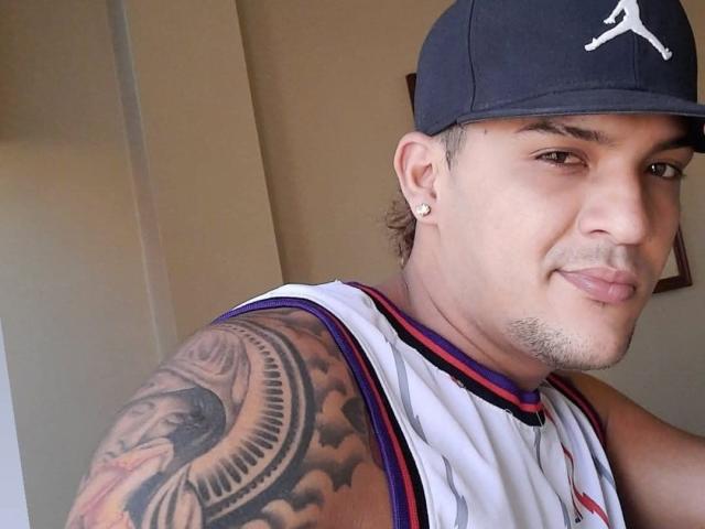 JorweenHot, wearing a baseball cap and white basketball jersey, shows off a detailed tattoo on his right arm, with a slight smile and a single ear piercing.